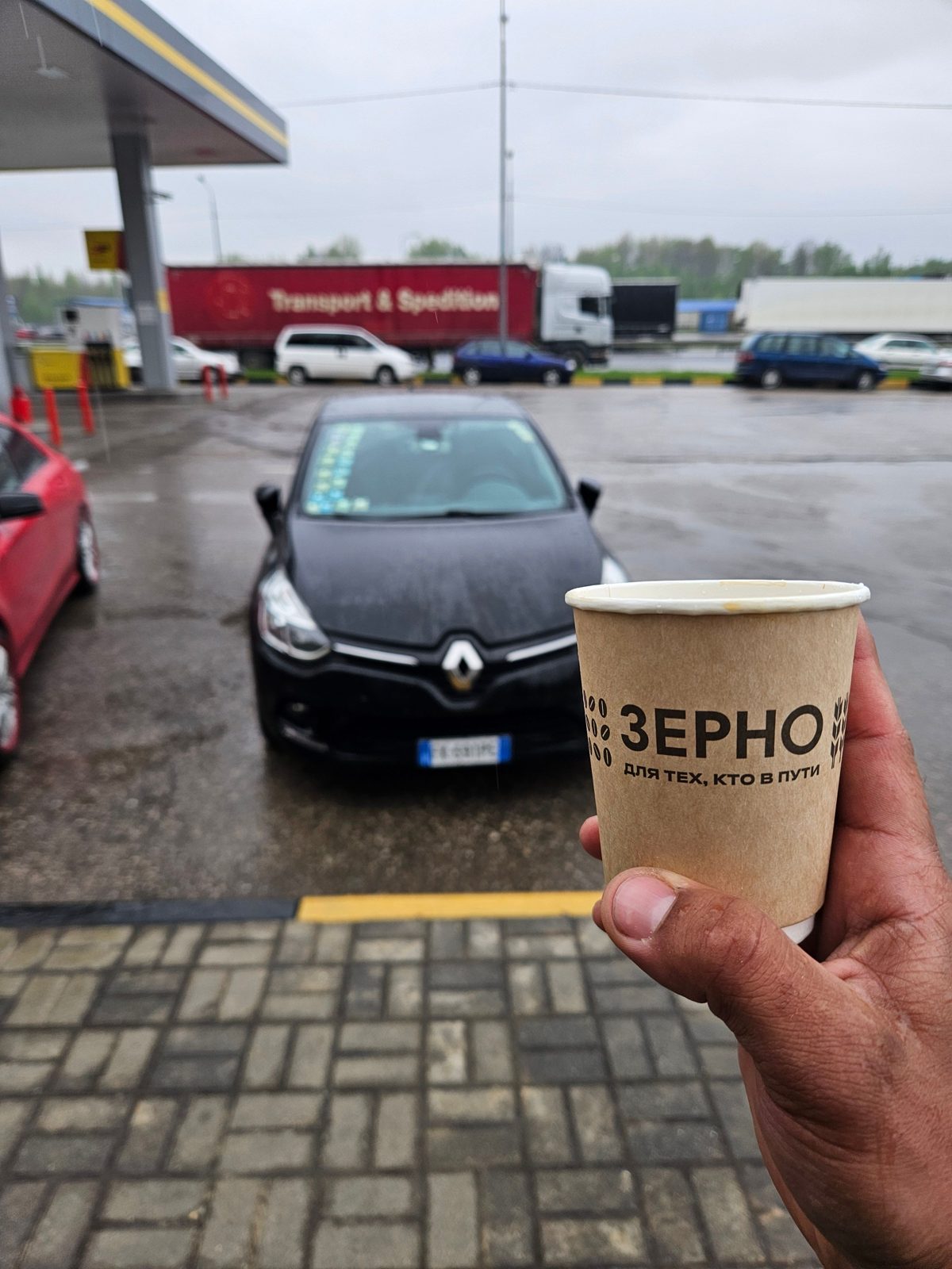 Una Renault Clio nera con targa italiana in una stazione di servizio russa e un caffè in mano